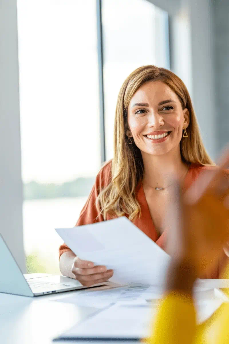 A woman with a bright smile sits at a table, focused on her laptop, embodying a moment of joy and concentration.