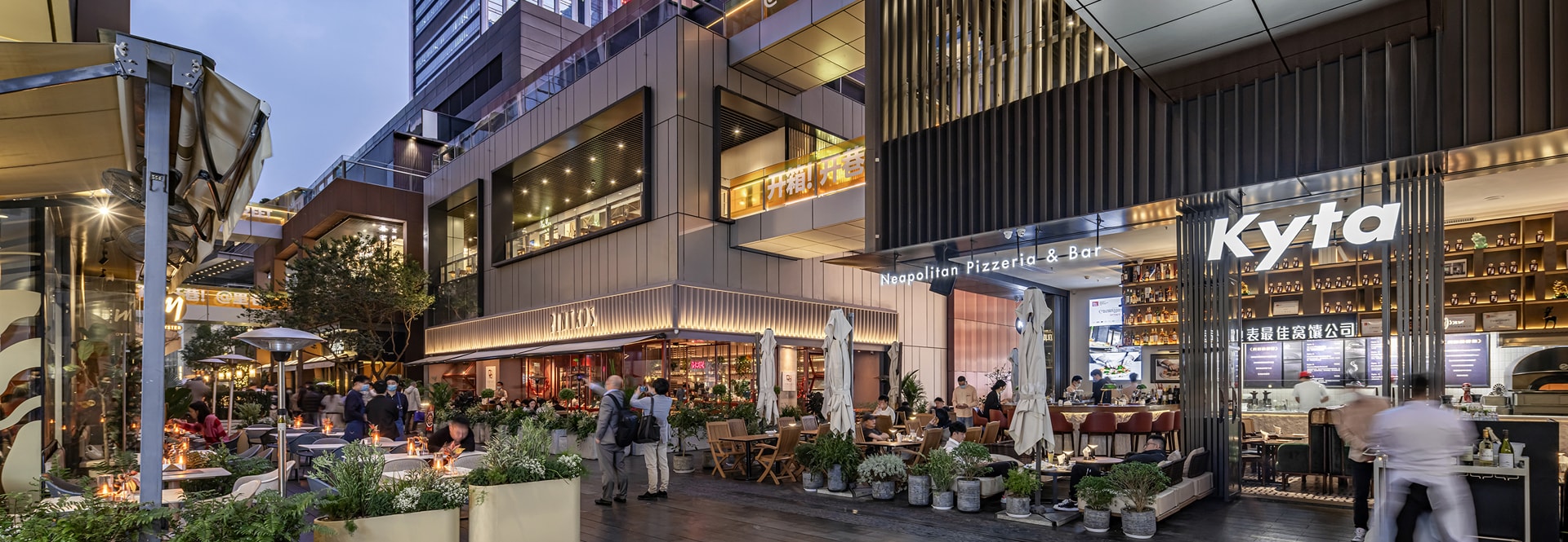 A bustling city street scene featuring people dining outdoors at tables, enjoying their meals in a lively atmosphere.