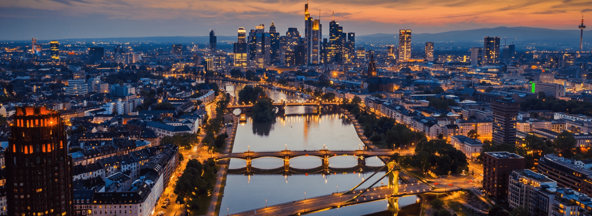 The Frankfurt skyline at sunset, featuring tall buildings outlined by warm hues of orange and pink in the sky.